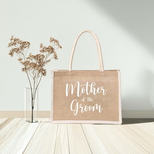 Pode incluir: Uma bolsa tote de juta bege com al&ccedil;as brancas e o texto "Mother of the Groom" impresso na frente.