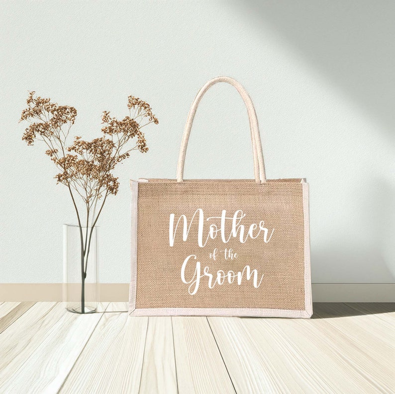 Pode incluir: Uma bolsa tote de juta bege com al&ccedil;as brancas e o texto "Mother of the Groom" impresso na frente.