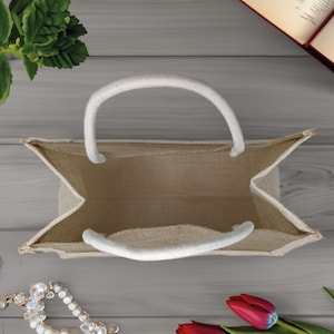 Pode incluir: Uma bolsa tote de juta bege com al&ccedil;as brancas. A bolsa est&aacute; vazia e aberta, mostrando o interior da bolsa.