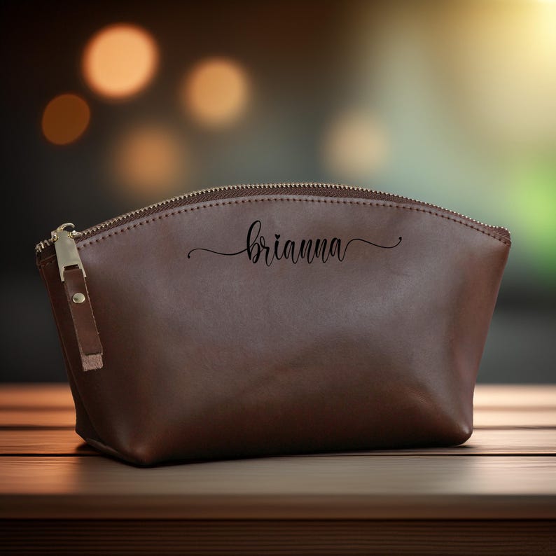 K&ouml;nnte beinhalten: Eine braune Kosmetiktasche aus Leder mit Rei&szlig;verschluss und kleinem Leder-Zuglasche. Der Name "brianna" ist in Schreibschrift auf die Vorderseite der Tasche geschrieben. Die Tasche steht auf einer Holzoberfl&auml;che.