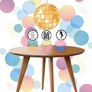 May include: Three cupcakes with different decorations on top of a wooden table. The cupcakes are decorated with frosting and sprinkles. The cupcake on the left has a black and white circle topper with the text "A lot going on at the moment". The cupcake in the middle has a black and white circle topper with the text "In my dance era". The cupcake on the right has a black and white circle topper with a silhouette of a woman dancing and a star.