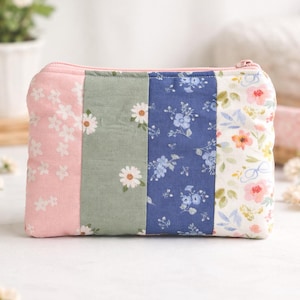 May include: A patchwork zippered pouch with a pink zipper and a floral pattern. The pouch is divided into vertical panels of pink with white flowers, green with daisies, blue with small flowers, and white with pink and orange flowers.