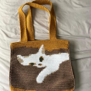 Può includere: Borsa tote all'uncinetto fatta a mano con un design a forma di gatto. La borsa ha un corpo marrone con una grafica di gatto bianco, finiture e cinghie gialle. La borsa è realizzata in filato ed è progettata per trasportare oggetti.