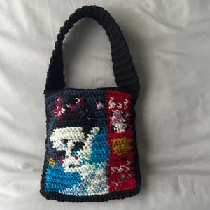 May include: A small, crocheted bag with a black strap and a colorful, pixelated design featuring a skull and other abstract shapes.