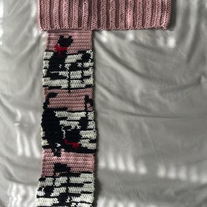 May include: A pink crocheted scarf with a black and white piano key pattern and a black cat design. The scarf is folded in half and laid flat on a white surface.