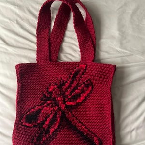 May include: A handmade, red crochet tote bag featuring a dragonfly design in black and red. The bag has two matching red handles and is made of a textured yarn. The dragonfly is centered on the front of the bag.