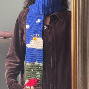 Könnte beinhalten: Ein blauer Häkel-Schal mit einem weißen Wolken-, gelben Sonnen- und grünem Gras-Design. Der Schal hat auch ein rotes und braunes Haus.