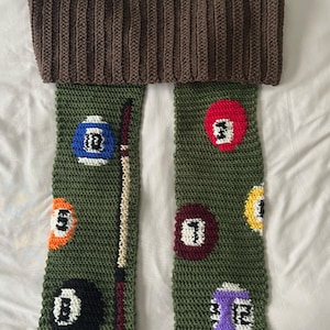 May include: A green and brown crocheted scarf with a pool ball design. The scarf features a brown crocheted band at the top and has a variety of colored pool balls with numbers on them.