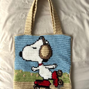 May include: Crocheted tote bag featuring Snoopy on a red skateboard against a blue sky and green grass background. The bag has beige handles and trim, with a light blue background. The Snoopy design is in white, brown, and black.