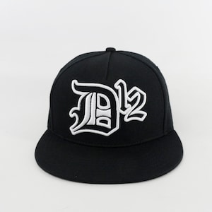 Op de afbeelding: Een zwarte, getailleerde baseballpet met een wit geborduurd "D12" logo in een gotisch lettertype.