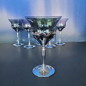 May include: Five silver-colored stemmed glasses with a decorative design. The glasses are arranged in a row, with the center glass in focus.