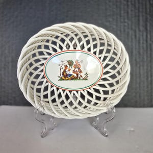 May include: A white, woven ceramic basket with an oval, central design. The design features a colorful illustration of two people in traditional clothing. The basket is displayed on a clear stand, showcasing its intricate, openwork design.
