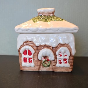 May include: A ceramic Christmas-themed jar shaped like a gingerbread house. The house has a white snow-covered roof and brick-like walls. Red-framed windows and a wreath adorn the front. A chimney and green garland add festive details.