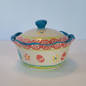 May include: A white ceramic bowl with a blue lid and a scalloped rim. The bowl is decorated with pink, yellow, and green floral and egg designs.