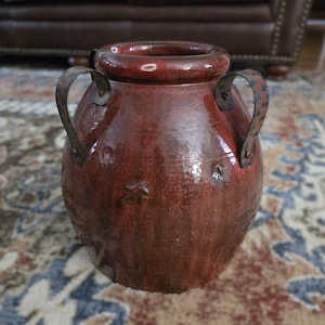 May include: A rustic, reddish-brown ceramic vase with two metal handles. The vase has a wide, rounded body and a narrow opening. The metal handles are attached to the sides of the vase with rivets. The vase is likely used for decorative purposes.