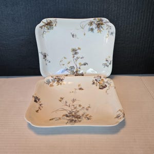 May include: Two square white ceramic plates featuring a delicate floral pattern in shades of blue, yellow, and brown. The plates have rounded corners and a smooth, glossy finish, with the floral designs scattered across the surface and edges.