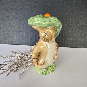 May include: A ceramic figurine of a rabbit wearing a green hat with an orange top. The rabbit is light brown with a white chest and is standing on a green base. The figurine is a decorative item.