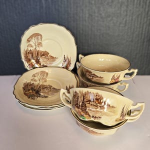 May include: A set of cream-colored ceramic soup bowls and saucers. Each piece features a brown landscape scene with trees and buildings. The saucers have a scalloped edge and the bowls have two handles.