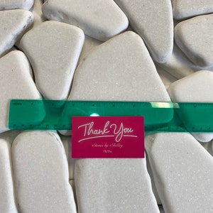 May include: A collection of smooth, white river stones of varying sizes. A green ruler measures the stones, and a pink card with "Thank You" in script is visible. The stones are ideal for crafts or home decor.