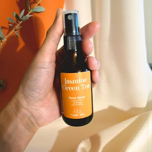 May include: A brown glass room spray bottle with a black spray nozzle and a bright orange label. The label reads "Jasmine Green Tea Room Spray 2 fl oz" in white text. The bottle is held in a hand.