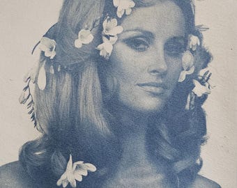 Può includere: Ritratto in tonalità blu di una persona con lunghi capelli mossi adornati da fiori bianchi. I fiori sono disposti tra i capelli, creando un aspetto naturale ed etereo. L'immagine ha una messa a fuoco morbida e un'estetica classica, vintage.