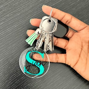May include: A clear acrylic keychain with the name "Sian" in black script and a teal "S" is attached to a silver keyring with keys and a mint green tassel. The keychain has glitter accents.
