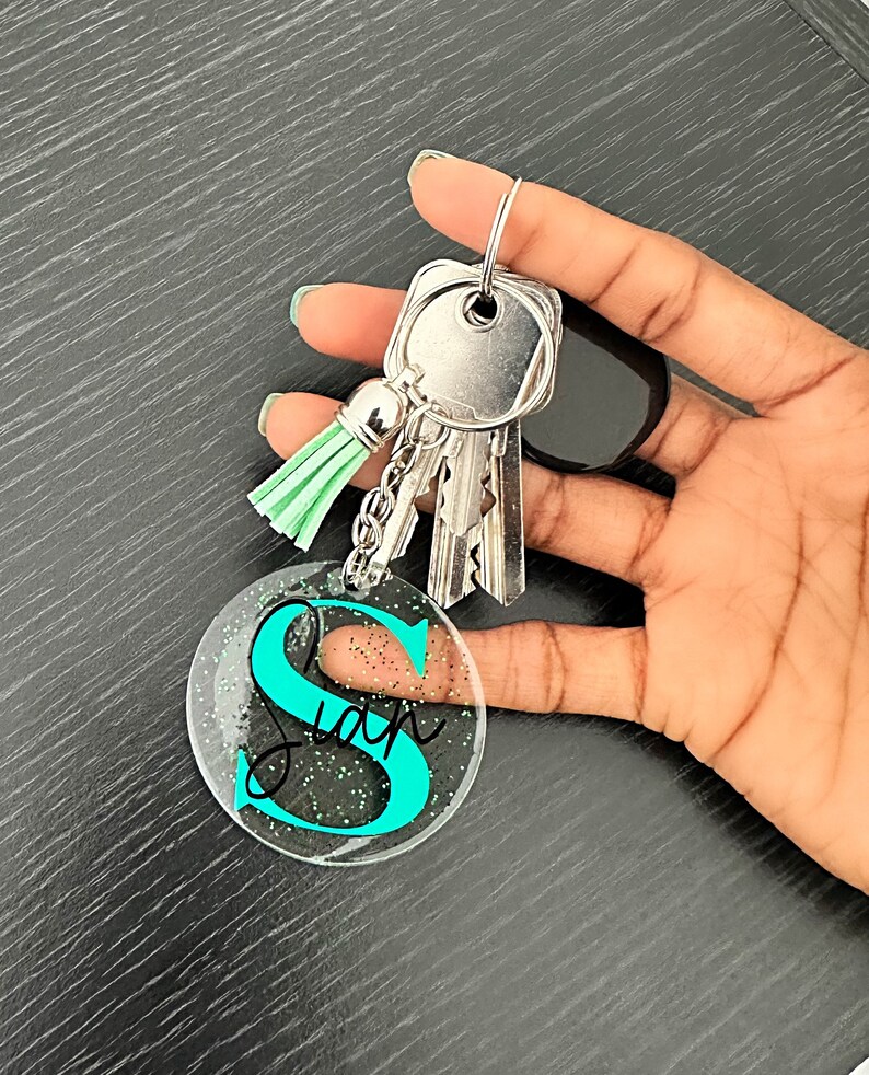 May include: A clear acrylic keychain with the name "Sian" in black script and a teal "S" is attached to a silver keyring with keys and a mint green tassel. The keychain has glitter accents.
