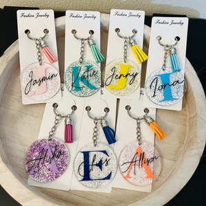 May include: A collection of personalized keychains with glitter accents. Each clear acrylic keychain features a different initial and name in various colors, with a matching tassel. The keychains are displayed on white cards that read "Fashion Jewelry".