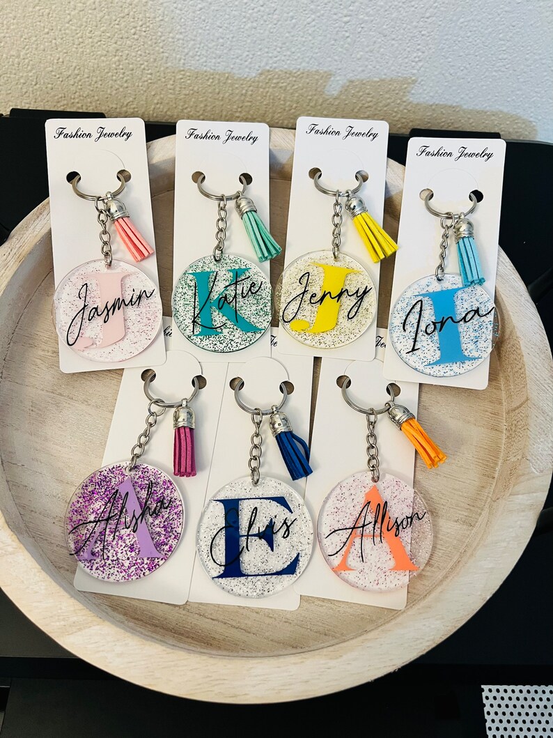 May include: A collection of personalized keychains with glitter accents. Each clear acrylic keychain features a different initial and name in various colors, with a matching tassel. The keychains are displayed on white cards that read "Fashion Jewelry".