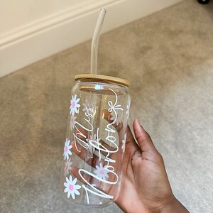 Pode incluir: Um copo de vidro transparente com tampa de bambu e canudo de vidro transparente. O copo &eacute; decorado com margaridas brancas com centros rosa e o texto cursivo "Miss Arlong".