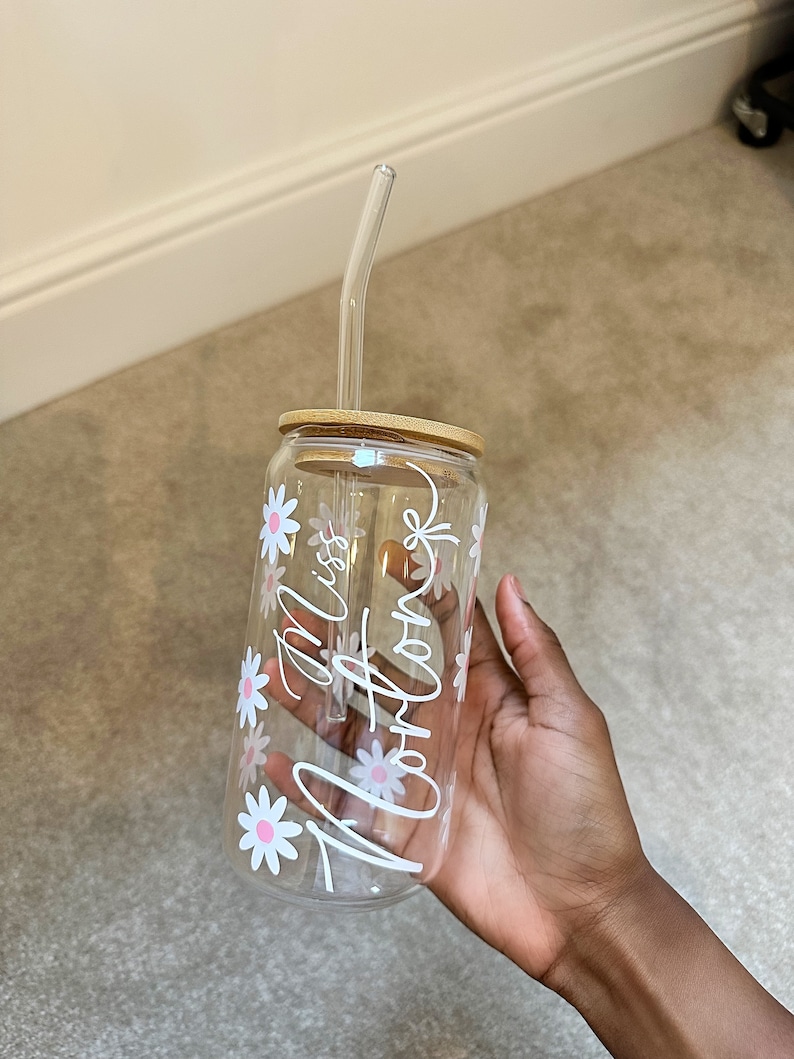 Pode incluir: Um copo de vidro transparente com tampa de bambu e canudo de vidro transparente. O copo &eacute; decorado com margaridas brancas com centros rosa e o texto cursivo "Miss Arlong".