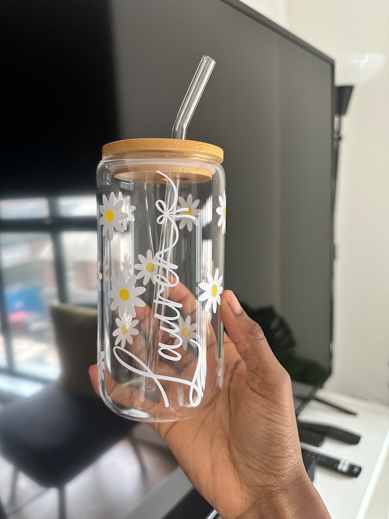 Pode incluir: Um copo de vidro transparente com tampa de bambu e canudo de vidro. O copo &eacute; decorado com margaridas brancas e o nome "Lauren" em escrita branca. O copo &eacute; segurado em uma m&atilde;o.