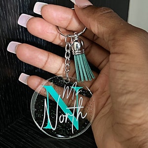 May include: A clear acrylic keychain with the name "Ms. North" in white script and a teal "N". The keychain has a silver keyring, chain, and a teal tassel. The background has black glitter.