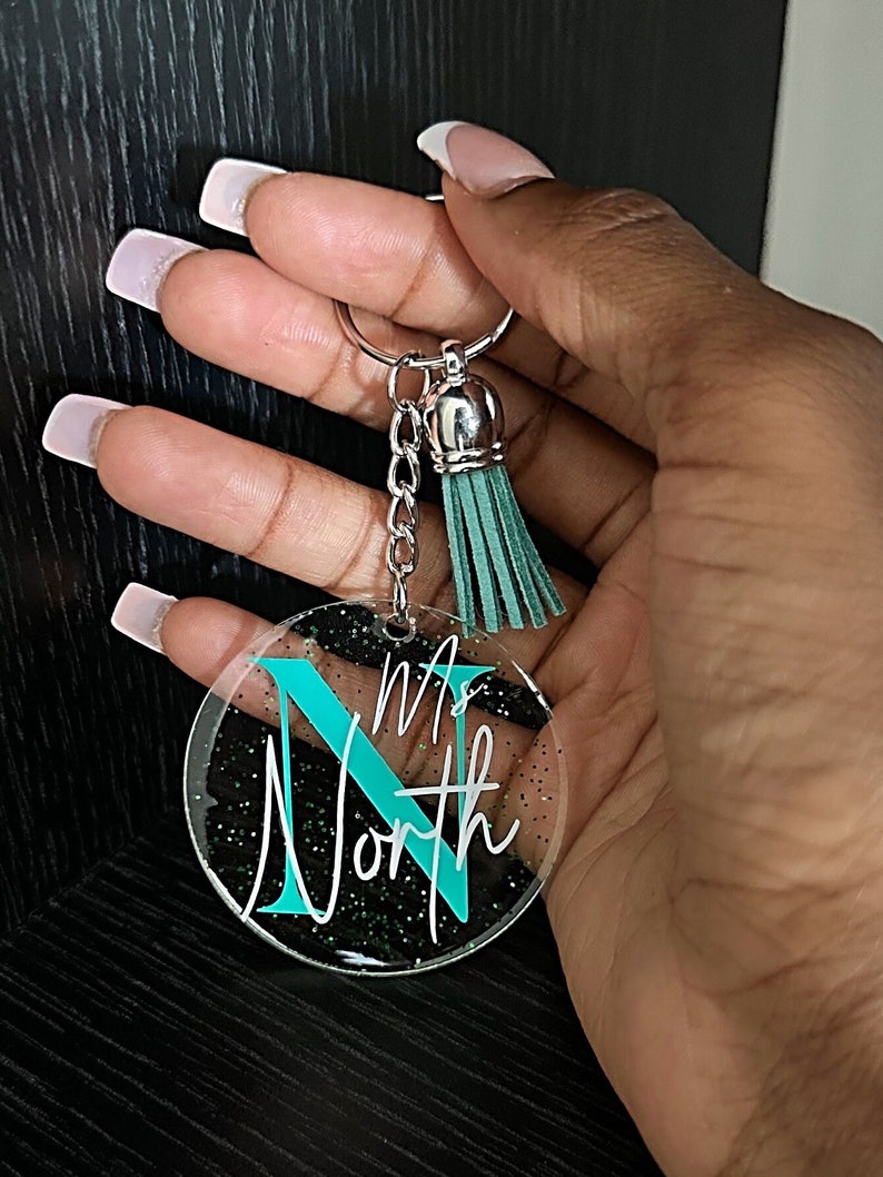 May include: A clear acrylic keychain with the name "Ms. North" in white script and a teal "N". The keychain has a silver keyring, chain, and a teal tassel. The background has black glitter.
