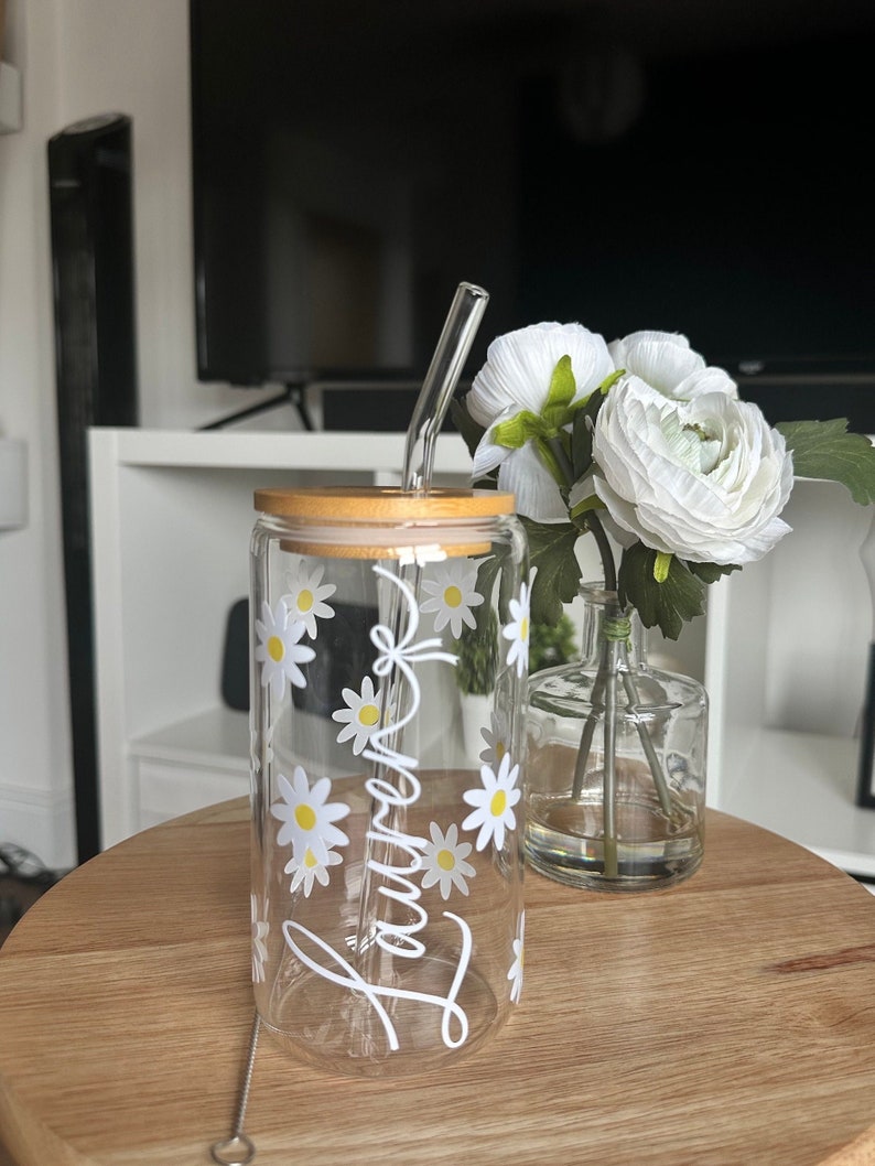 Pode incluir: Um copo de vidro transparente com tampa de bambu e canudo de vidro. O copo &eacute; decorado com flores de margarida brancas e o nome "Lauren" em it&aacute;lico. Um limpador de canudo de a&ccedil;o inoxid&aacute;vel &eacute; vis&iacute;vel. Um vaso de flores brancas est&aacute; por perto.