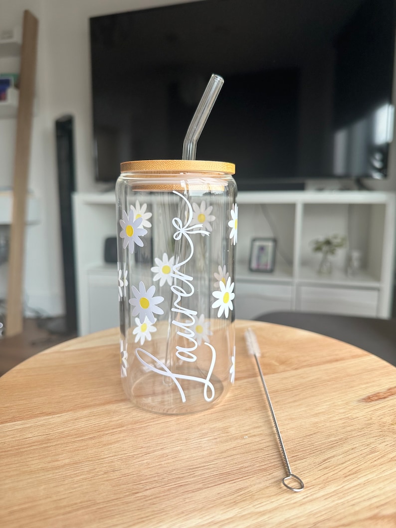 Pode incluir: Um copo de vidro transparente com tampa de bambu e canudo de vidro transparente. O copo &eacute; decorado com margaridas brancas e o nome "Lauren" em escrita branca. Um limpador de canudos de a&ccedil;o inoxid&aacute;vel est&aacute; ao lado do copo.