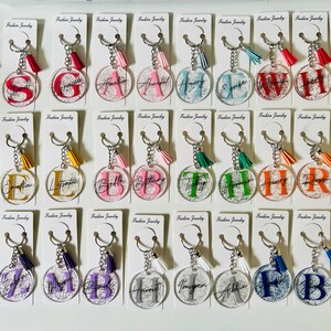 May include: A collection of clear acrylic keychains with various initials in different colors. Each keychain has a silver chain, a tassel, and a card that says "Fashion Jewelry". The initials are in a variety of fonts and colors.