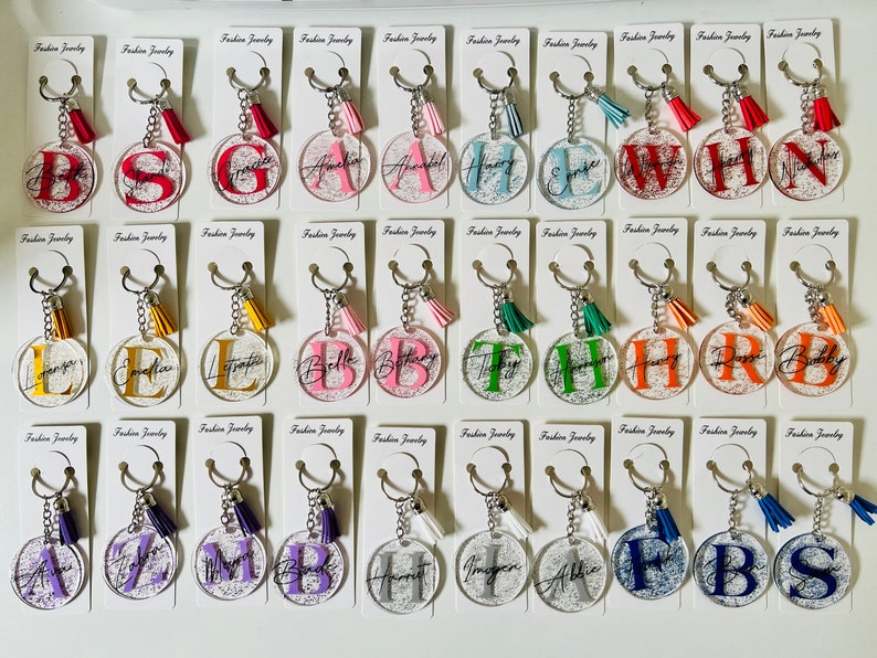 May include: A collection of clear acrylic keychains with various initials in different colors. Each keychain has a silver chain, a tassel, and a card that says "Fashion Jewelry". The initials are in a variety of fonts and colors.