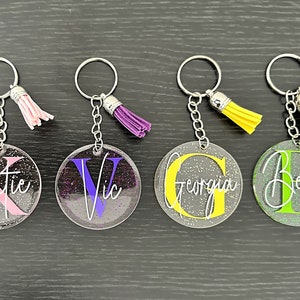 May include: Four personalized keychains with glitter accents. Each keychain features a clear, round acrylic disc with a name and initial in various colors: pink, purple, yellow, and green. Each keychain has a matching tassel.