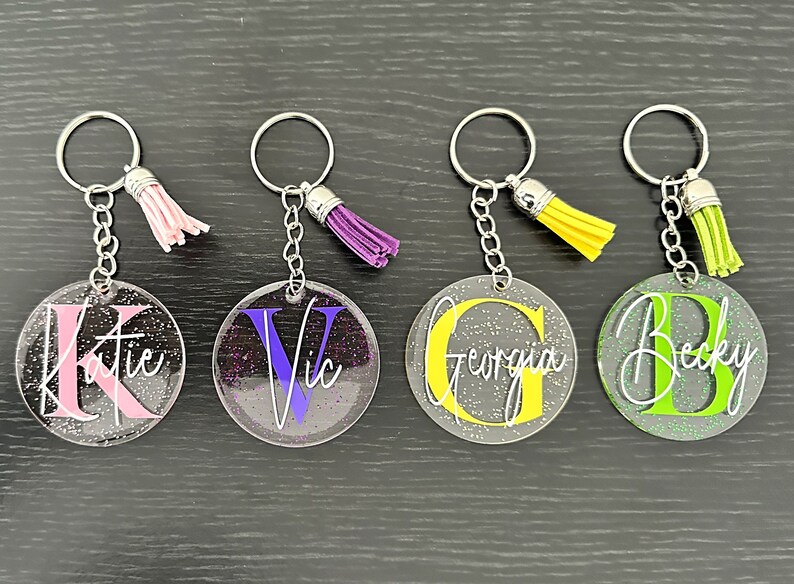 May include: Four personalized keychains with glitter accents. Each keychain features a clear, round acrylic disc with a name and initial in various colors: pink, purple, yellow, and green. Each keychain has a matching tassel.