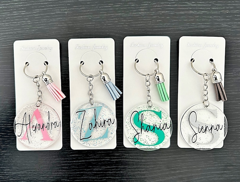 May include: Four personalized keychains with silver hardware, each featuring a clear, round acrylic charm with a name and initial. The keychains have colorful tassels in pink, blue, green, and brown. The names are Alexandra, Zahira, Shania, and Sienna.