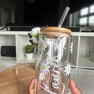 Pode incluir: Um copo de vidro transparente com tampa de bambu e canudo de vidro. O copo &eacute; decorado com flores de margarida brancas e o nome "Lauren" em escrita branca. Uma escova de limpeza est&aacute; ao lado.