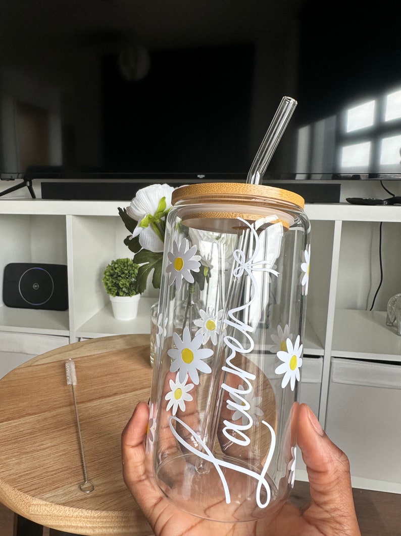 Pode incluir: Um copo de vidro transparente com tampa de bambu e canudo de vidro. O copo &eacute; decorado com flores de margarida brancas e o nome "Lauren" em escrita branca. Uma escova de limpeza est&aacute; ao lado.