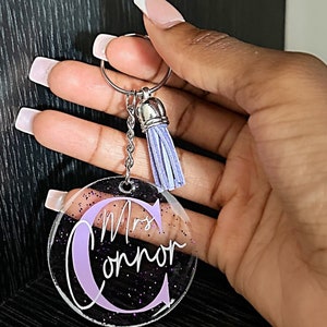 May include: Clear acrylic keychain with the name "Mrs. Connor" in white script and a large lavender letter "C". The keychain has a silver keyring and chain, and a lavender tassel. The background has purple glitter.