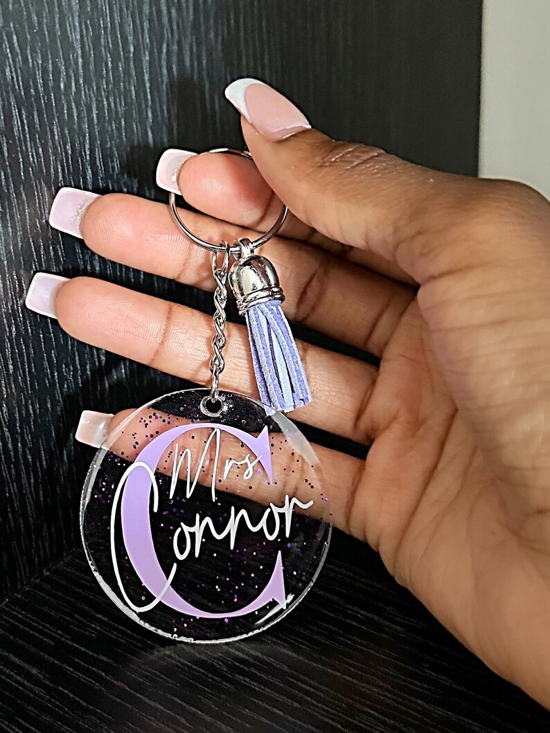 May include: Clear acrylic keychain with the name "Mrs. Connor" in white script and a large lavender letter "C". The keychain has a silver keyring and chain, and a lavender tassel. The background has purple glitter.
