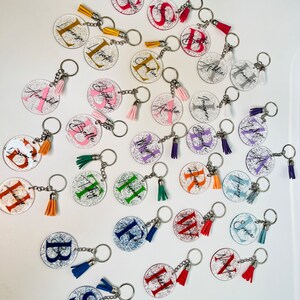May include: A collection of personalized keychains with glitter accents. Each clear, circular keychain features a letter or name in various colors, paired with a matching tassel. The keychains are attached to silver key rings.