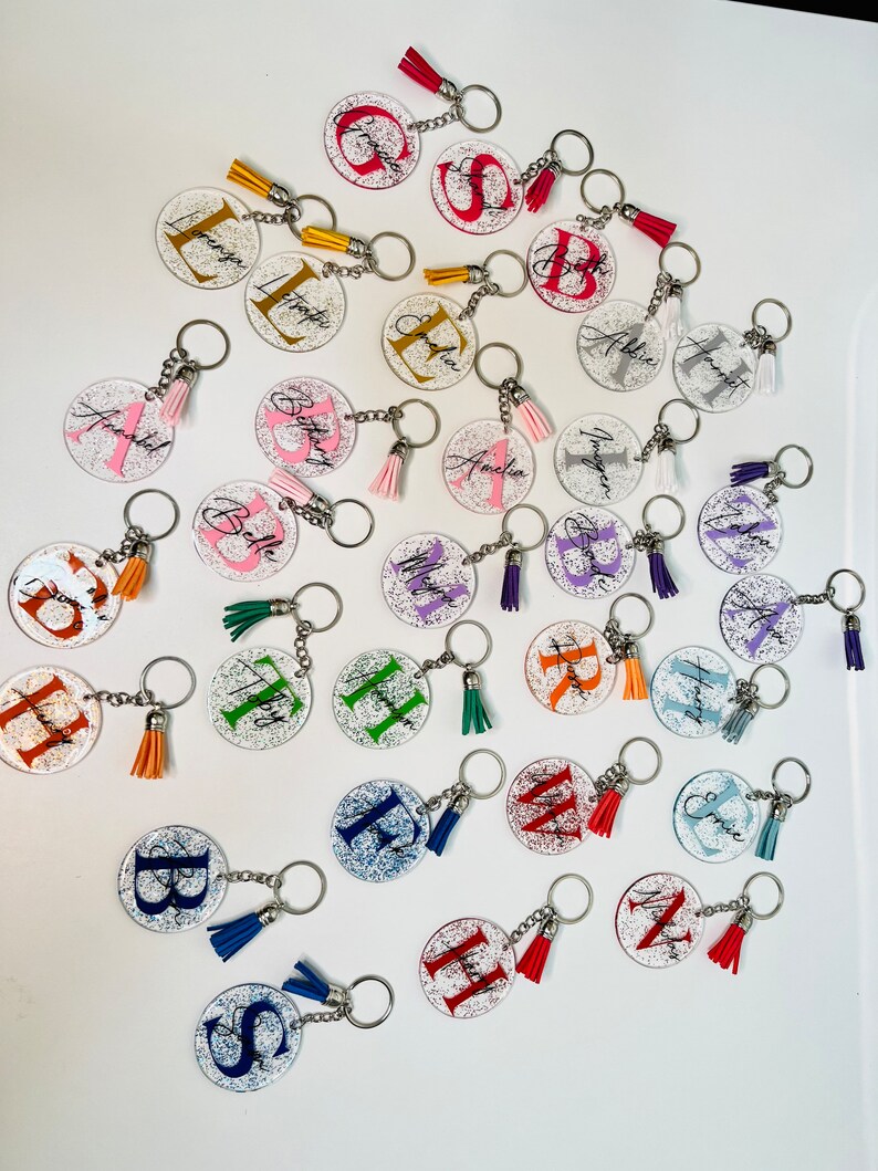 May include: A collection of personalized keychains with glitter accents. Each clear, circular keychain features a letter or name in various colors, paired with a matching tassel. The keychains are attached to silver key rings.