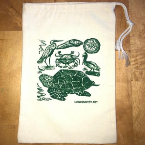 May include: A white canvas drawstring bag with a green print of a sea turtle, a crab, a bird, a seashell, and a sand dollar. The text "LOWCOUNTRY ART" is printed on the bag.