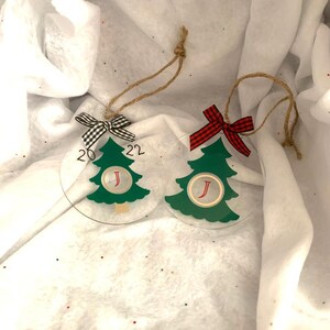 May include: Two clear Christmas tree ornaments with green trees, the year 2022, and the letter "J". One has a black and white checkered bow, the other a red and black checkered bow. Both have twine hangers.