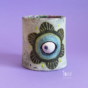 Puede incluir: Una taza de cerámica blanca con un diseño de ojo verde, azul y blanco. La taza tiene una superficie texturizada y está decorada con puntos amarillos. El texto "TOGU CERAMICS" es visible en la parte inferior de la imagen.